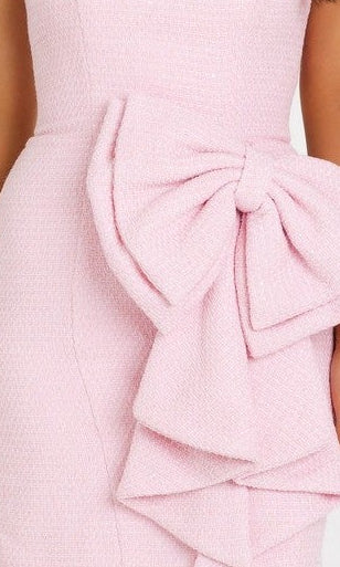 A woman stands against a white background, smiling in the Ava Presley 42917 short homecoming dress—a light pink, strapless tweed mini with a statement bow detail on the skirt—paired with nude heels.